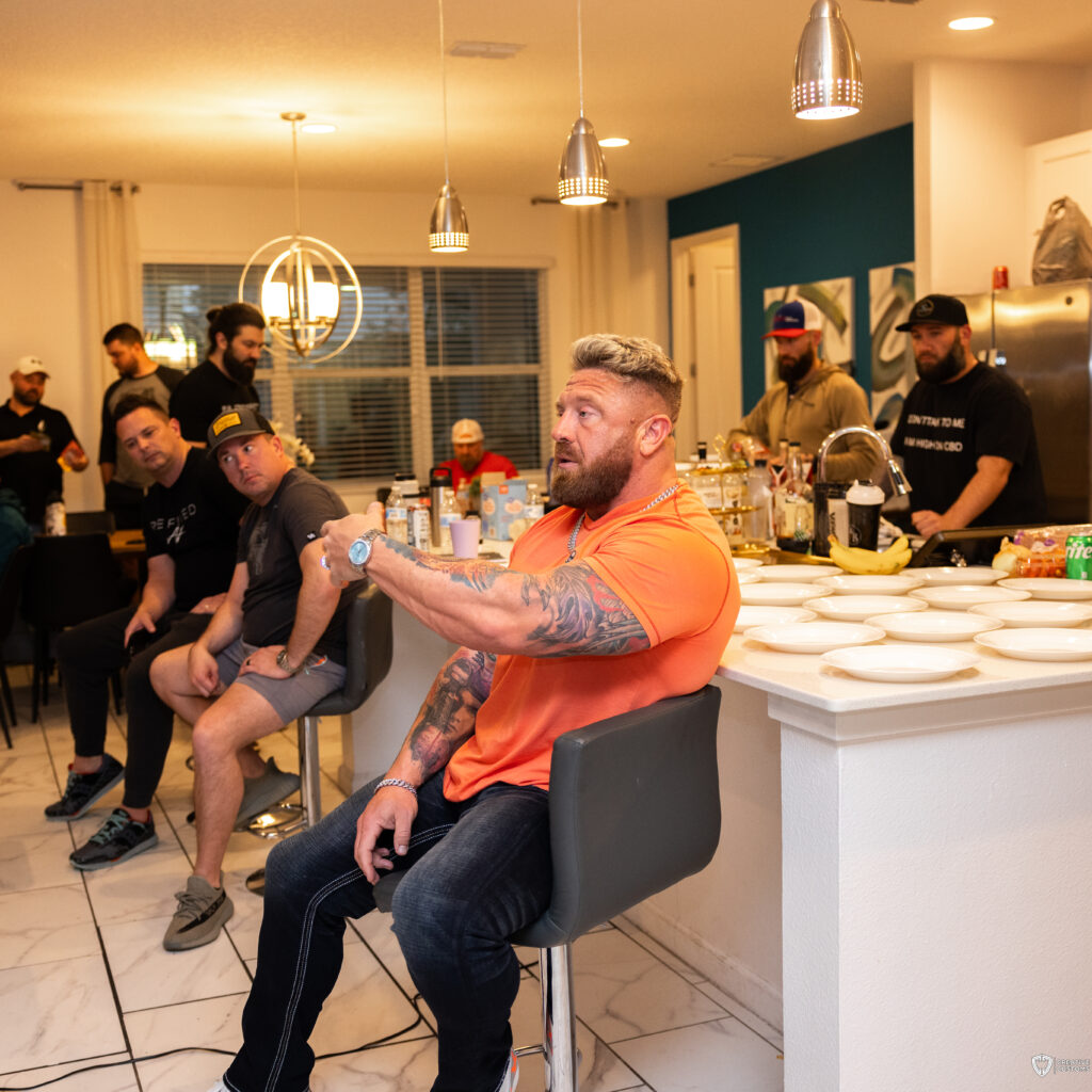 A muscular, tattooed man in an orange shirt passionately speaks while sitting at a kitchen island, engaging with a group of men attentively listening in the background at an Uncaged Men's Accountability Group event.