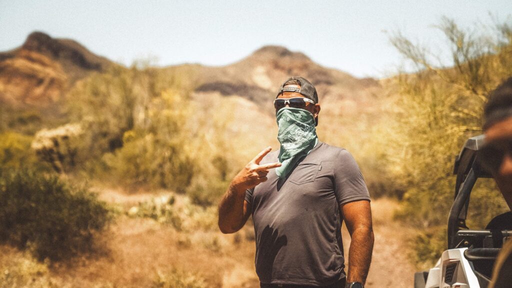 A man wearing sunglasses and a bandana over his face gives a peace sign in the middle of the desert during an Uncaged Men’s Accountability Group retreat, embracing authenticity and adventure.