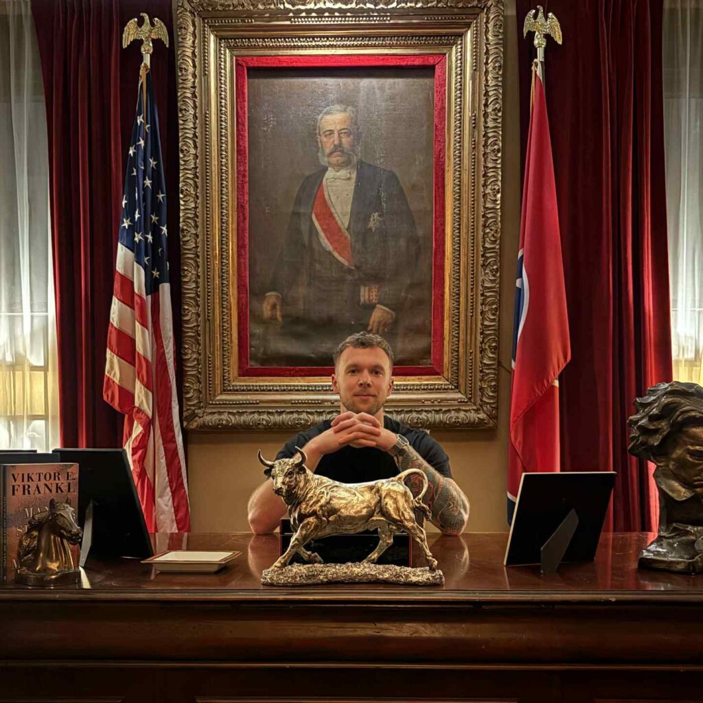 A man sitting confidently at a grand, historic desk with American and state flags behind him, surrounded by statues and classic decor, embodying leadership and legacy.